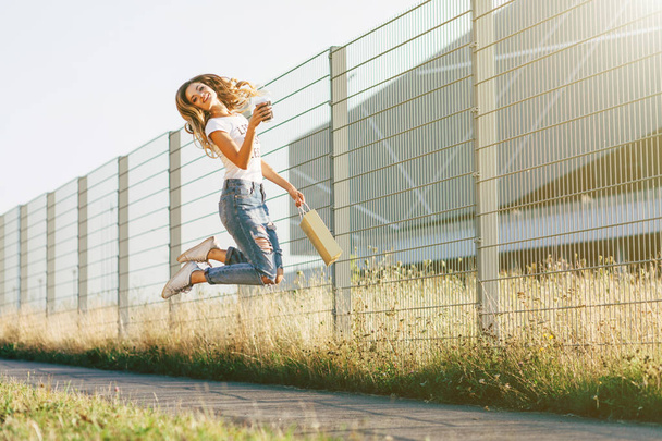 Retrato de una hermosa joven en camiseta blanca caminando cerca de una cerca de metal con una taza de café en las manos
 - Foto, imagen