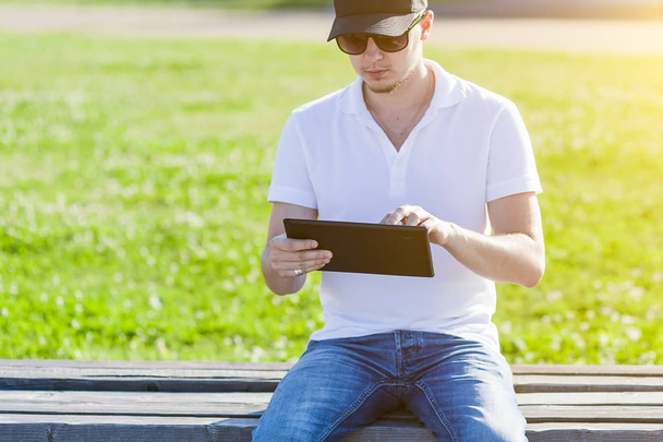Young adult man using pc tablet outdoor in city park on bench. Man reading at park. - Foto, afbeelding