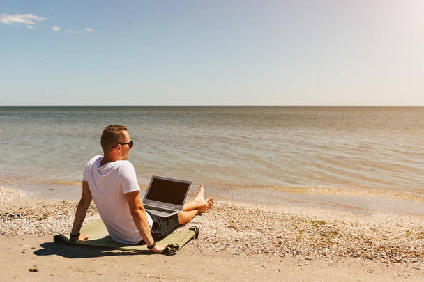 Joven en una camiseta blanca se sienta en la playa con un ordenador portátil en el fondo del mar
 - Foto, imagen