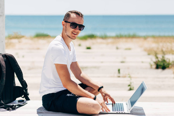 Jonge man in een wit T-shirt zit op het strand met een laptop op de achtergrond van de zee - Foto, afbeelding