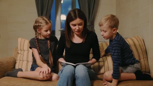 Mutter sitzt mit Tochter und kleinem Sohn auf der Couch und liest ihnen eine Geschichte vor, während sie auf der Couch im Wohnzimmer sitzen. - Filmmaterial, Video