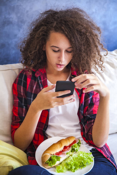 Curly hair teen girl using mibile phone and eating sandwich - Фото, изображение