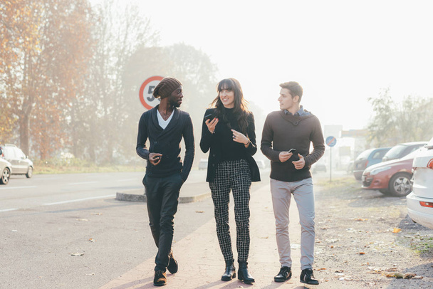 Tres amigos de mujeres y hombres multiétnicos caminando al aire libre en la ciudad luz de fondo, charlando y sosteniendo el teléfono inteligente
 - Foto, imagen