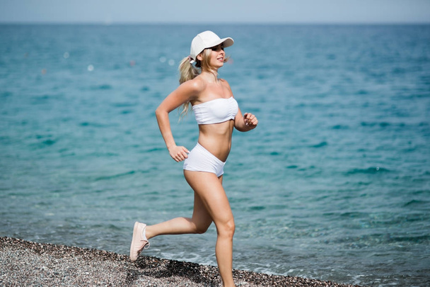 Beautiful young blonde woman in white sportswear is running on beach - Photo, Image
