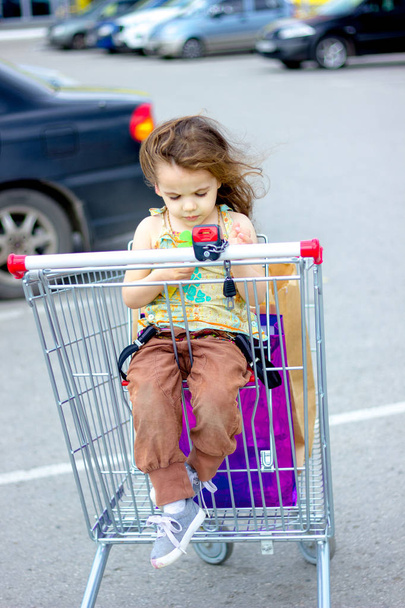 Little kid girl sitting in a shopping cart near mall. - Fotó, kép