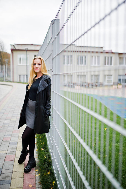 Blonde fashionable girl in long black leather coat against iron  - Photo, image