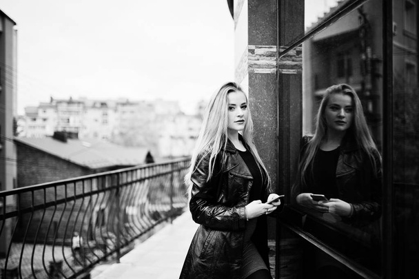 Blonde fashionable girl in long black leather coat posed against - Foto, Bild