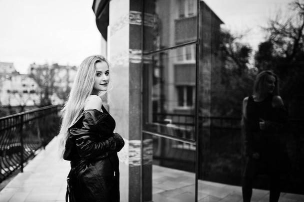 Blonde fashionable girl in long black leather coat posed against - Фото, изображение