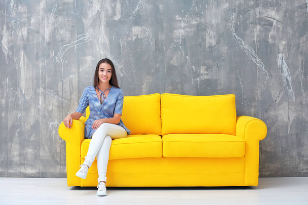 Young woman sitting on yellow sofa near grey wall - 写真・画像