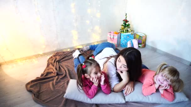 Cute female and two little girls children, posing for camera and smiling, lying on cushions, on floor in room with garland and fir tree in evening. - Footage, Video