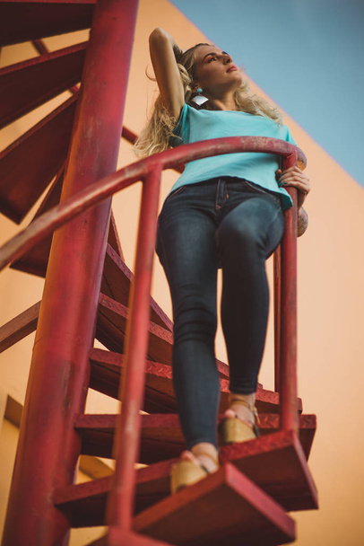 Beautiful leggy blonde is standing on a red spiral staircase and looking into the distance. On the background of orange walls and sky. Modern street fashion - Photo, image