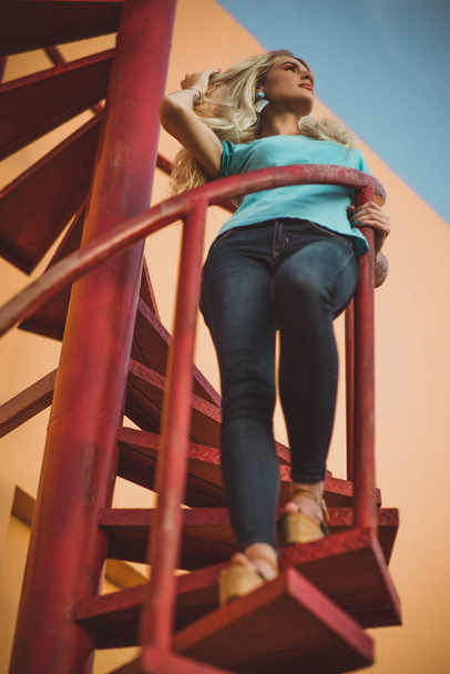 Beautiful leggy blonde is standing on a red spiral staircase and looking into the distance. On the background of orange walls and sky. Modern street fashion - 写真・画像