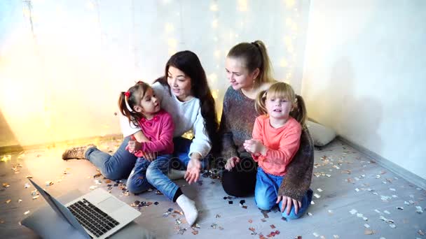 older women spend time together with younger girl children and sisters using laptop and sitting on floor on background of wall with garland in room. - Footage, Video