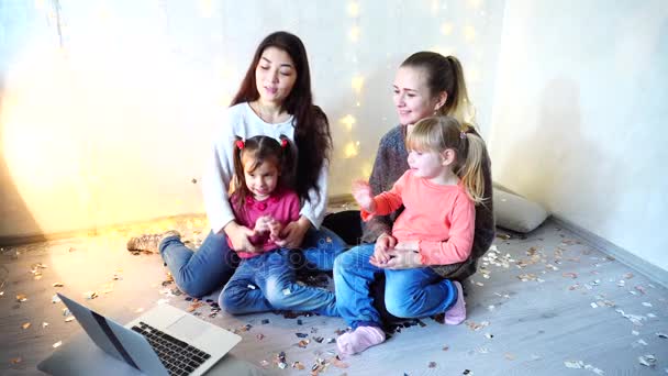 Deux femmes plus âgées passent du temps avec les jeunes filles enfants et sœurs en utilisant un ordinateur portable et assis sur le sol sur le fond du mur avec guirlande dans la chambre
. - Séquence, vidéo
