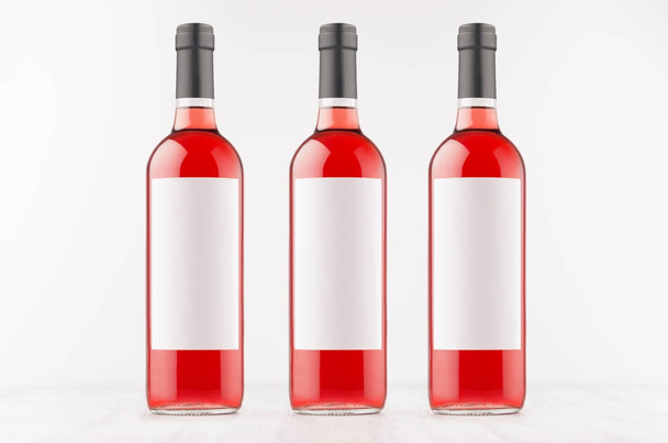 Three rose wine bottles with blank white labels on white wooden board, mock up. - Foto, afbeelding