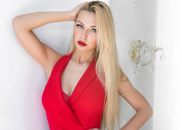 Fashion image of young smiling beautiful woman in dress.Close-up portrait of young elegant blonde woman in red dress and red lips touching her long blonde hair. Fashion indoor shot - Foto, Imagen