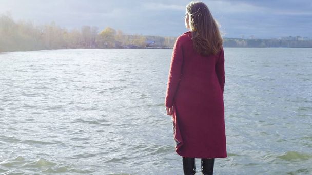 Young girl with her hair stands near the water in the cold season. Back view. Young woman standing near a lake - Photo, Image