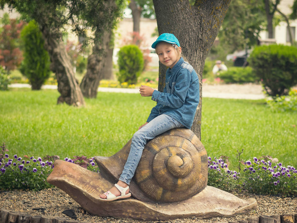 公園でカタツムリの像にまたがって座っている青い野球帽で美しい少女. - 写真・画像