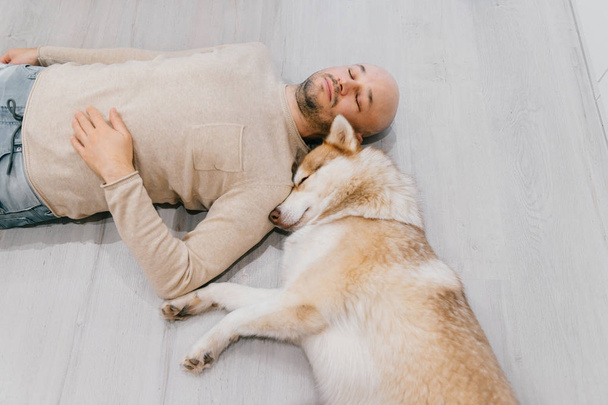 Adult bald man with husky puppy sleeping on floor. Owner with pet together at home. Kind and soulful emotions. Lovely dog resting with young male. Guy with beloved domestic animal hugging each other. - Photo, Image