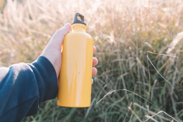 Yellow bottle in hand. Travel concept. Hand holding yellow flask outdoors. - Фото, зображення