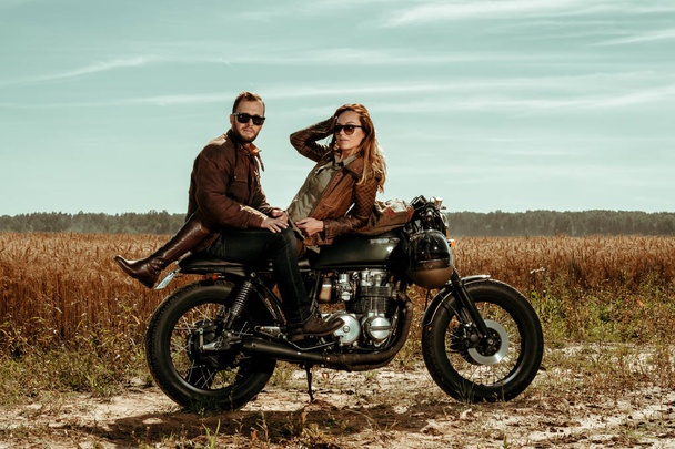 Young, stylish cafe racer couple on the vintage custom motorcycles in a field. - Photo, Image