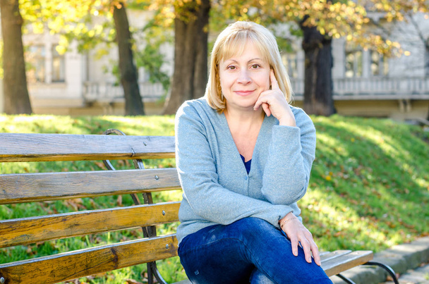   Блондинка середнього віку сидить на лавці в парку, дивлячись на камеру на осінньому весняному фоні
.  - Фото, зображення