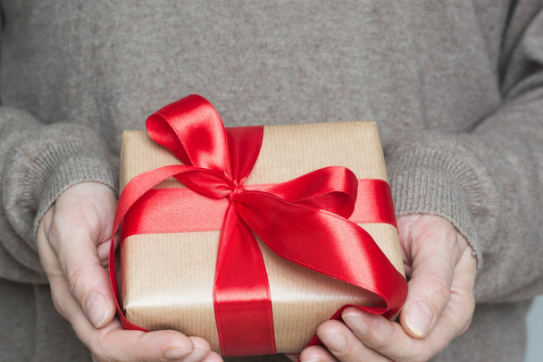 Female hands in woolen beige sweater holding beige box wrapped in craft paper with red ribbon. Close up. - Photo, Image