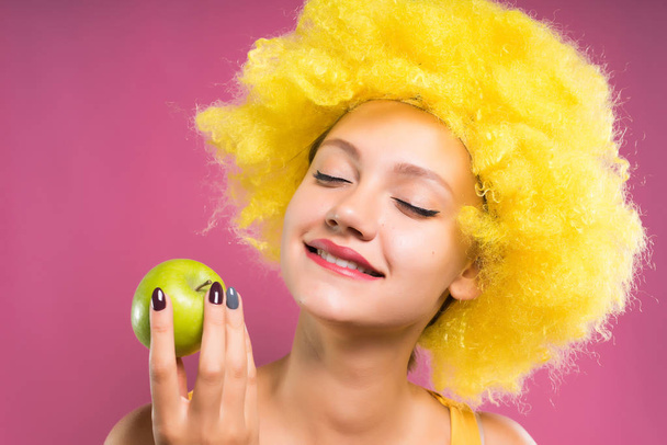 funny girl in yellow clown wig holding an apple in her hand, smiling - Fotó, kép