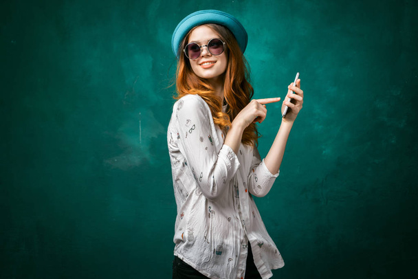 charming stylish girl in a blue beret and sunglasses is holding a smartphone and points at it with her finger, smiling - Photo, Image