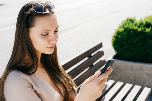 beautiful attractive girl in a beige dress sits on a wooden bench, looks into her smartphone - Foto, Imagen
