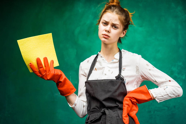 sad young girl in a black apron, wearing orange rubber gloves holding a yellow rag - Fotografie, Obrázek