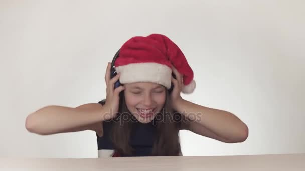 Krásná zlobivá holka teenager v Santa Claus klobouk poslouchá hudbu ve sluchátkách a zpívá spolu na bílém pozadí stopáže videa. - Záběry, video