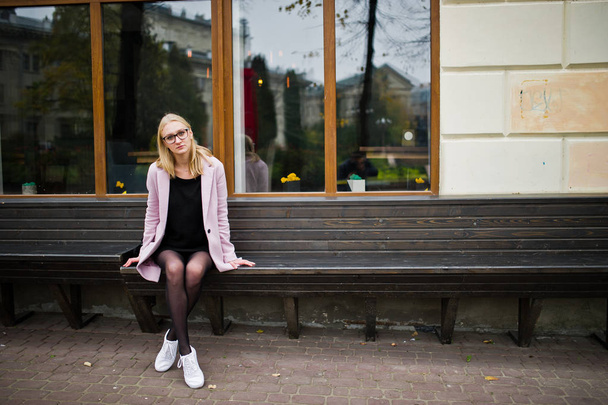 Blonde girl at glasses and pink coat, black tunic sitting on ben - Photo, image