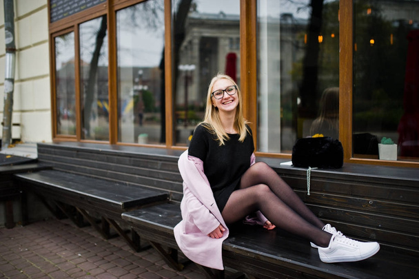 Blonde girl at glasses and pink coat, black tunic sitting on ben - Photo, Image