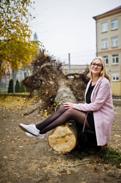 Blonde girl at glasses and pink coat, black tunic sitting on cut - Photo, image