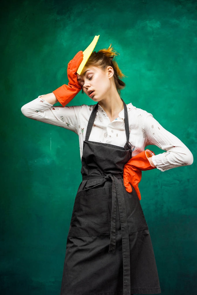 a young girl in a black protective apron, rubber gloves wipes sweat from her forehead because she is tired - Foto, Imagen