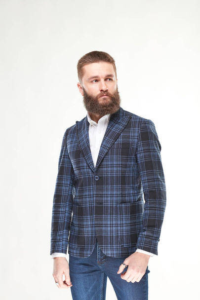 Attractive young sexy serious man model with beard posing in blue and black jacket, white shirt and dark blue pants in light studio  - Foto, afbeelding
