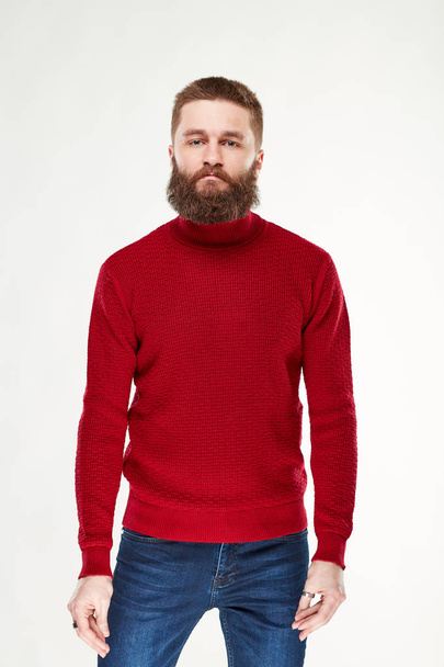 Attractive young sexy man model with beard and dark hair posing in red sweater and dark blue pants in light studio  - Фото, изображение