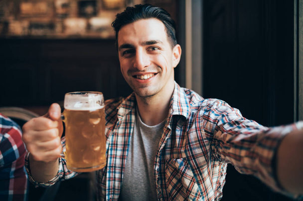 Strzał z podekscytowany mężczyzna młody krzyczy, szczęśliwie biorąc selfie ze szklanką piwa relaks w lokalnym pubie - Zdjęcie, obraz