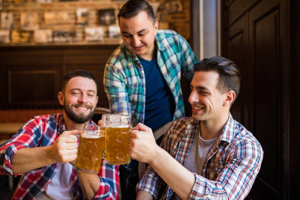 Троє молодих чоловіків в повсякденному одязі посміхаються і стискають келихи пива разом, сидячи в пабі
 - Фото, зображення