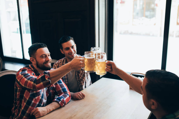 Drie gelukkige jongemannen in casual kleding zijn glimlachend en rinkelende glazen bier samen zittend in pub - Foto, afbeelding