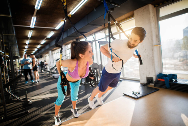 beautiful sporty woman exercising with TRX system in gym, muscular personal trainer assisting her - Fotografie, Obrázek