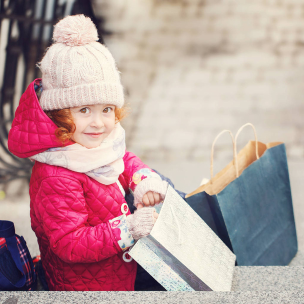 Cute little girl with shoppings bags. Shopping and sale concept. Child is wearing a warm hat, jacket and scarf. - Foto, imagen