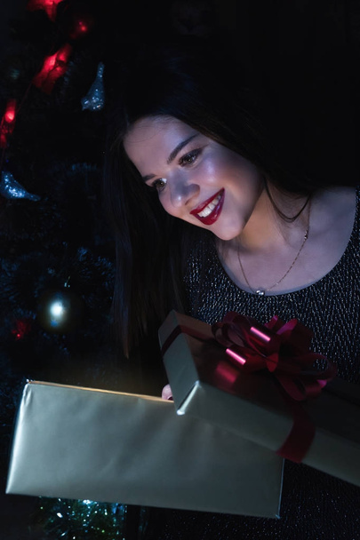Дівчина тримає подарунок, сидячи під ялинкою
 - Фото, зображення