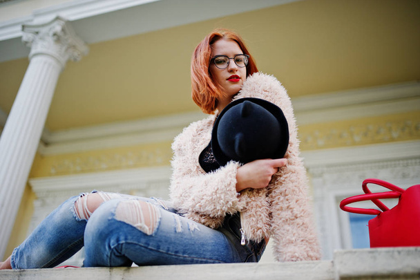 Red haired girl in glasses and hat with red handbag posed near v - Фото, зображення