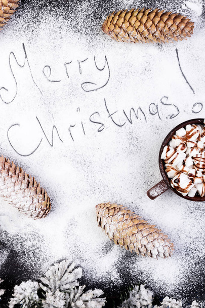 Feliz Navidad Texto hecho a mano en la nieve. Comida de Navidad Sweet Marshmallow con Chocolate en Brown Cup sobre fondo negro. Piso Lay
.  - Foto, imagen