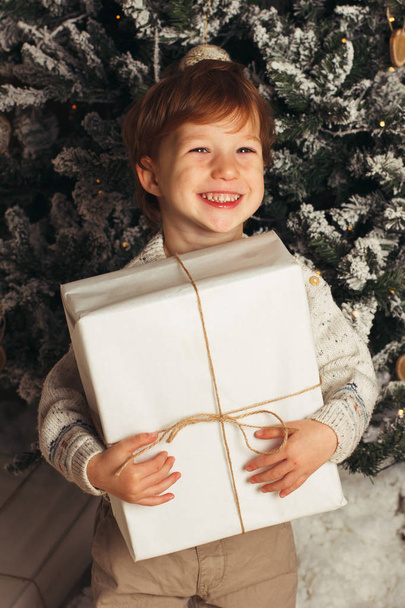 Christmas portrait of a young boy cozy atmosphere around the Christmas tree. Cute toddler holding white gift box. Vertical photo, copy cpace - Fotografie, Obrázek