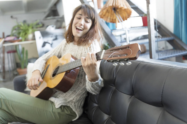 Beautiful woman sitting on a sofa and  playing guitar - Foto, imagen
