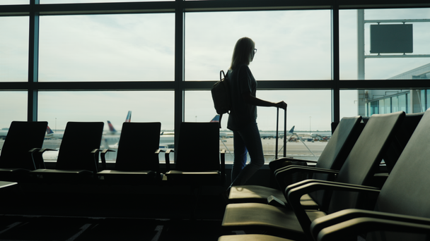 Eine Geschäftsfrau mit Reisetasche blickt in einem großen Fenster auf die Flugzeuge. im Flughafenterminal stehen - Filmmaterial, Video
