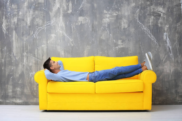 Young man lying on yellow sofa near grey wall - Foto, Imagen